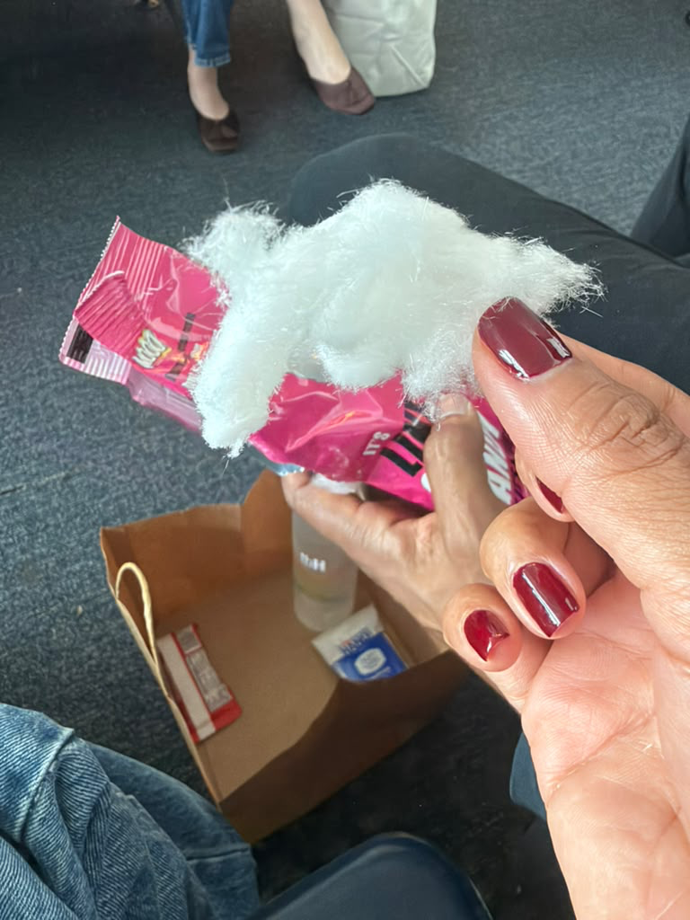 Hand holding pink-packaged white fluffy food resembling cotton candy, with brown paper bag and shoes in background.
